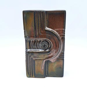 May include: A ceramic vase with a geometric design in shades of brown, green, and black. The vase has a unique, abstract shape with a circular opening at the top.