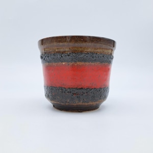 Peut inclure: Un pot en céramique à surface texturée. Le pot présente des bandes horizontales marron, noires et rouges. Les bandes supérieure et inférieure sont marron, avec une large bande rouge au milieu. Le pot est de forme cylindrique.