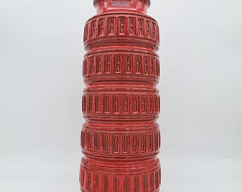 May include: A tall, cylindrical ceramic vase in a vibrant red glaze. The exterior features five stacked, repeating horizontal bands with a textured, geometric pattern of vertical indentations. A small, rectangular label is visible near the rim.