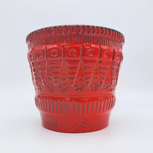 Könnte beinhalten: Ein leuchtend roter Keramik-Pflanztopf mit glänzender Oberfläche und strukturiertem Reliefmuster. Das Design umfasst vertikale Dreiecksformen, kreisförmige Akzente und kleine Blumenmotive am oberen Teil sowie einen gerippten Rand und Boden.
