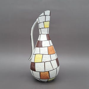 May include: A decorative ceramic jug with a curved handle. The jug features a geometric design of white, brown, yellow, and orange squares outlined in black. The jug has a slender neck and a wide base.
