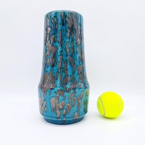 Op de afbeelding: Een blauwe keramische vaas met een zilveren en blauwe gespikkelde glazuur. De vaas is hoog en smal, met een ronde bodem en een uitlopende bovenkant. Hij staat op een wit oppervlak naast een gele tennisbal.