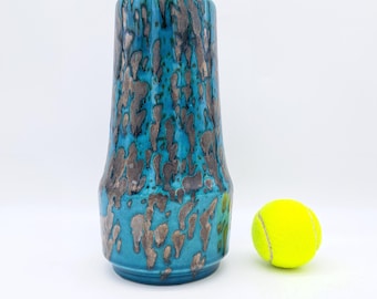 Könnte beinhalten: Eine blaue Keramikvase mit einem silbernen und blauen gesprenkelten Überzug. Die Vase ist hoch und schmal, mit einem abgerundeten Boden und einem ausgestellten oberen Rand. Sie steht auf einer weißen Oberfläche neben einem gelben Tennisball.