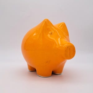 Könnte beinhalten: Ein glänzendes, orangefarbenes Sparschwein aus Keramik. Das Schwein hat einen abgerundeten Körper, kleine Ohren und eine Schnauze. Es steht auf vier kleinen Füßen. Das Sparschwein ist ein klassisches Design zum Sparen von Geld.