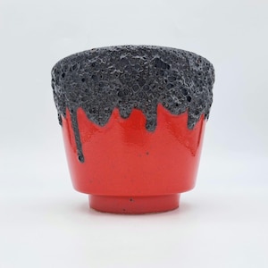 Peut inclure: Une tasse en céramique avec une base rouge vif et un émail noir poreux et texturé qui coule depuis le bord. Le récipient présente une forme conique avec une base surélevée, soulignant le contraste entre la surface rouge lisse et le bord sombre.
