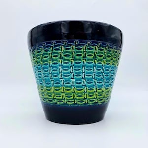 May include: A ceramic planter with a glossy black rim and base. The body features a repeating pattern of green and blue ovals, creating a textured, eye-catching design. The planter is suitable for indoor or outdoor use.