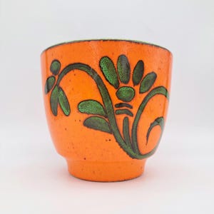 Op de afbeelding: Een feloranje keramische beker met een donkergroen bloemmotief en een textuurafwerking. De beker heeft een afgerond lichaam dat taps toeloopt naar een kleinere ronde basis, wat zorgt voor een opvallend kleurcontrast en een handgeschilderd uiterlijk.