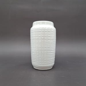 Puede incluir: Un jarrón de cerámica blanco con un patrón texturizado similar al tejido. El jarrón cilíndrico tiene una parte superior ligeramente ensanchada y una base redondeada. La superficie presenta una serie de pequeñas hendiduras espaciadas, agregando interés visual. Fondo gris oscuro.