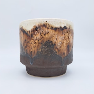 Pode incluir: Um vaso de cerâmica com um design em camadas. A parte superior é branco sujo, passando para um padrão castanho e castanho escuro, e terminando com uma base castanha escura. O vaso tem uma forma cilíndrica com uma base ligeiramente elevada.