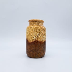 Puede incluir: Un jarrón cilíndrico de cerámica con superficie texturizada. La parte superior es de color beige moteado, que pasa a una base marrón oscuro. El jarrón tiene un borde ligeramente ensanchado.