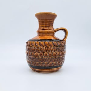 Może przedstawiać: Brązowy ceramiczny wazon z uchwytem. Wazon ma teksturowany, wzorzysty design z ciemnym pasem w pobliżu podstawy i góry. Wazon ma około 15 cm wysokości.