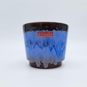 May include: A ceramic planter with a blue and brown glaze. The planter has a cylindrical shape with a slightly flared base. The top rim is dark brown, transitioning to blue with a dripped effect. The brand name "scheurich" is on a red sticker.