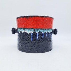 Peut inclure: Un vase cylindrique en céramique avec un extérieur bleu foncé texturé. La section supérieure est rouge, et une bande de blanc et de vert sépare les sections rouge et bleue. Deux petites poignées noires sont de chaque côté.