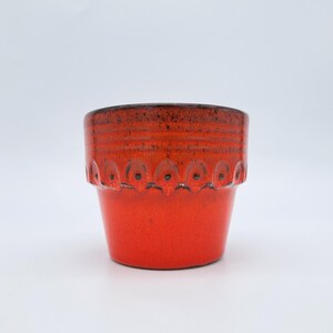 May include: A vibrant, small, ceramic planter in a bright red hue. The top rim is a darker shade of red, and a decorative band of repeating circular patterns encircles the middle. The planter is approximately 4 inches tall and 4 inches in diameter.