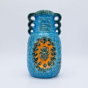 Puede incluir: Un jarrón de cerámica azul con una superficie texturizada y un diseño decorativo naranja y verde. El jarrón tiene tres juegos de tres asas circulares en la parte superior. El jarrón mide aproximadamente 25 cm de alto.