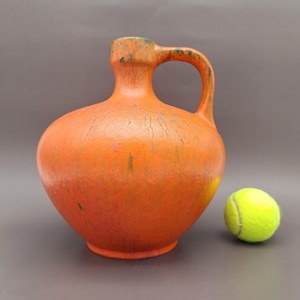 Può includere: Brocca in ceramica arancione con manico, accanto a una pallina da tennis gialla. La brocca ha un corpo arrotondato e un collo stretto. La brocca è alta circa 25 cm.