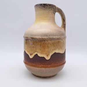 Puede incluir: Un jarrón de cerámica marrón y amarillo con una boca ancha y un asa. El jarrón tiene un efecto de glaseado de goteo y una banda marrón alrededor del medio.