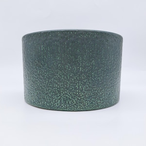 May include: A dark teal ceramic planter, cylindrical in shape, with a textured, speckled surface. The planter has a wide opening and a rounded base. The color is a deep, muted green with lighter green speckles.