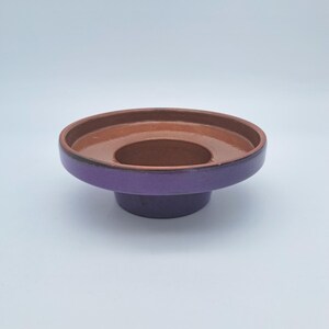 Pode incluir: Uma tigela de cerâmica circular com uma borda larga e plana e uma base central elevada. O exterior da tigela tem um acabamento em esmalte roxo profundo, enquanto o interior e a borda superior apresentam uma superfície de argila castanho-avermelhada.