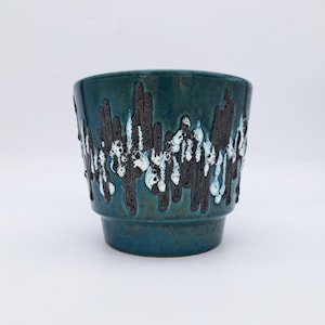 Op de afbeelding: Een blauwgroene keramische plantenpot met een taps toelopende voet en een brede rand. De buitenkant heeft een gestructureerd, verticaal druippatroon in donkerbruin en wit glazuur dat contrasteert met het gladde, diepblauwgroene oppervlak.