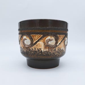 Peut inclure: Un pot de fleurs en céramique marron foncé avec un motif texturé. Le pot présente une bande de détails texturés avec un motif tourbillonnant répété dans des tons de marron, de beige et de blanc. La base et le bord sont marron foncé brillant.