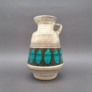 Pode incluir: Um vaso de cerâmica vintage com uma alça, apresentando um corpo texturizado bege e marrom. O vaso tem uma faixa de formas ovais turquesas em um fundo marrom escuro, adicionando um toque de cor ao design.
