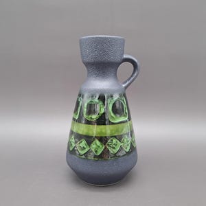 Puede incluir: Un jarrón de cerámica azul oscuro con un patrón geométrico verde y negro. El jarrón tiene una boca ancha y un asa.