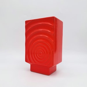 Può includere: Un vaso in ceramica rosso vivo, di forma rettangolare, con un motivo strutturato di cerchi concentrici su un lato. Il vaso ha una finitura lucida e una piccola base rettangolare. Un oggetto decorativo per la casa.