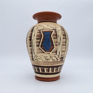 Può includere: Un vaso decorativo in ceramica con bordo e base marrone, caratterizzato da un motivo strutturato beige e marrone. Un disegno di vaso blu è centrato sulla parte anteriore. Il vaso ha un corpo largo e un collo stretto.