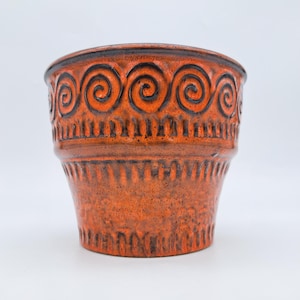 Peut inclure: Un cache-pot cylindrique en céramique arborant un vernis orange vif avec des accents brun foncé. La partie supérieure présente un motif répétitif de spirales noires, tandis que le corps inférieur est orné de rainures verticales texturées.