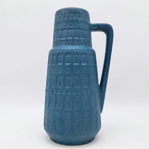 May include: A blue ceramic pitcher with a textured geometric pattern. The pitcher has a handle and a wide base, tapering to a narrower neck. The pitcher is approximately 12 inches tall.
