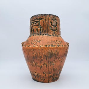 Puede incluir: Un jarrón de cerámica alto y texturizado en tonos marrón y naranja. El jarrón presenta diseños en relieve de un ciervo y formas abstractas cerca de la parte superior. Dos pequeñas asas están en los lados. El jarrón tiene una estética rústica y vintage.