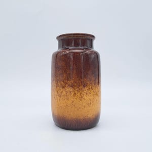 Puede incluir: Un jarrón de cerámica marrón con un cuerpo cilíndrico y un borde ligeramente ensanchado. El jarrón tiene un efecto degradado, que va del marrón oscuro en la parte superior a un marrón anaranjado claro y moteado en la base. El jarrón está sobre un fondo blanco.