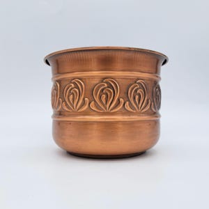 Op de afbeelding: Een cilindervormige koperen plantenbak met een gladde afwerking en een decoratieve band van in reliëf aangebrachte, herhalende krulpatronen rond het bovenste gedeelte. Het metaal heeft een warme, metaalachtige oranjebruine kleur.