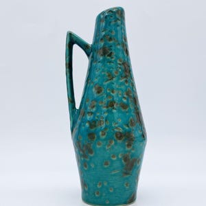 Op de afbeelding: Een hoge, turquoise keramische vaas met een handvat. De vaas heeft een gespikkeld bruin patroon en een unieke, hoekige vorm. Het handvat is een eenvoudige, open lus. De vaas is een decoratief item.