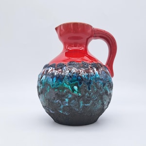 Puede incluir: Una jarra de cerámica roja con asa y base texturizada multicolor. La base presenta tonos de azul, verde y negro. La jarra es un objeto decorativo, probablemente para exhibición o servicio.