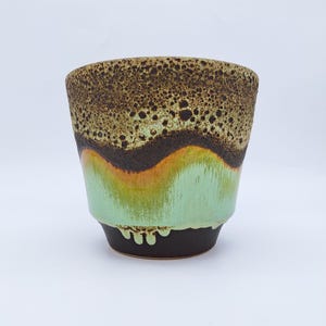 Puede incluir: Una taza de cerámica con una parte superior marrón texturizada, una banda marrón oscuro y una sección inferior verde claro y naranja. La base es marrón oscuro con un diseño de goteo. La taza tiene forma cilíndrica.