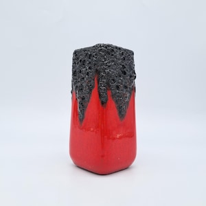 Puede incluir: Un jarrón de cerámica roja con una parte superior negra texturizada. El jarrón tiene forma rectangular con esquinas redondeadas. La parte superior negra tiene un aspecto poroso, similar a la lava, que contrasta con el cuerpo rojo liso y brillante. Una pieza decorativa para el hogar.