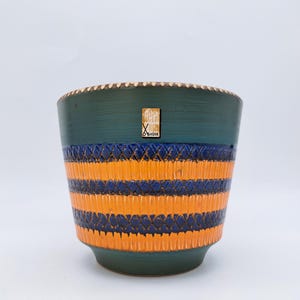 Puede incluir: Una maceta de cerámica con exterior verde azulado y rayas horizontales texturizadas en naranja y azul marino. El borde tiene un borde decorativo. Una pequeña etiqueta con texto está adherida al lateral. Adecuado para uso en interiores o exteriores.