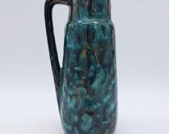 May include: A tall, ceramic vase with a handle, featuring a textured, dripped glaze in shades of teal, turquoise, and dark brown. The vase has a cylindrical body and a dark handle. The overall aesthetic is vintage and decorative.