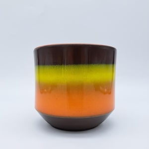 Könnte beinhalten: Ein zylindrischer Keramik-Pflanztopf mit Farbverlauf. Die oberen und unteren Bereiche sind dunkelbraun, in der Mitte geht es in Gelb und Orange über. Der Topf hat eine glatte, glänzende Oberfläche.