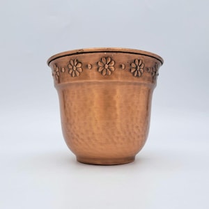 Puede incluir: Una maceta de metal color cobre con una textura martillada en el cuerpo y una banda decorativa alrededor del borde superior que presenta un patrón repetitivo de flores de margarita en relieve. El recipiente tiene una boca ancha y base cónica.