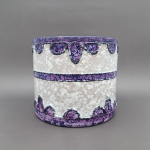 May include: A cylindrical ceramic vase with a textured white surface and purple accents. The vase features a band of purple around the middle and decorative scalloped edges in purple. The vase has a speckled appearance.