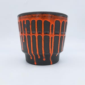 Op de afbeelding: Een matzwarte keramische beker met een feloranje glazuur dat vanaf de rand langs de zijkanten naar beneden druipt. De beker heeft een brede opening die taps toeloopt naar een smallere basis, wat een opvallend silhouet met een donkere textuur geeft.
