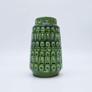 May include: A green ceramic vase with a geometric pattern of squares and circles. The vase has a cylindrical shape with a slightly flared base and a narrow neck. The glossy finish reflects light, highlighting the textured design.