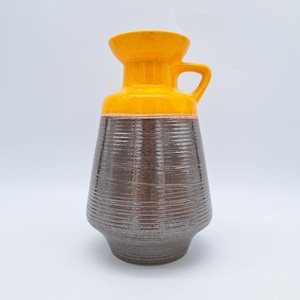 May include: A brown and yellow ceramic vase with a textured surface and a handle. The vase has a wide mouth and a narrow base.