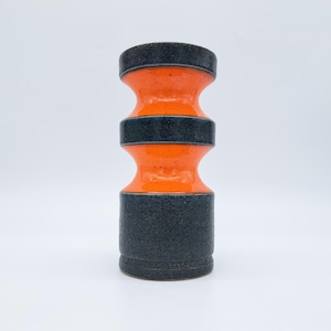 Puede incluir: Un jarrón de cerámica con una forma de reloj de arena única. El jarrón presenta una sección central de color naranja vibrante, contrastada por bandas de color gris oscuro texturizado en la parte superior, media e inferior. El jarrón tiene un acabado brillante y está sobre un fondo blanco.