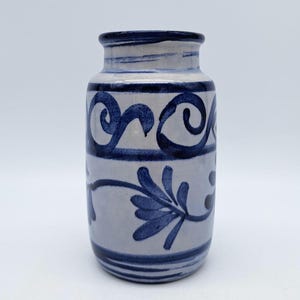 Puede incluir: Un pequeño jarrón de cerámica blanca con diseños florales y espirales azules. El jarrón tiene un cuerpo redondeado y un labio ligeramente acampanado.