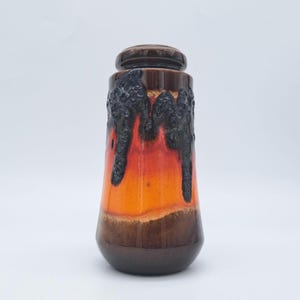 Puede incluir: Un jarrón de cerámica con tapa, con un degradado de color que va del marrón oscuro al naranja y al negro. El jarrón tiene un diseño texturizado, similar a la lava, en la parte superior. La base es de un tono marrón más oscuro.
