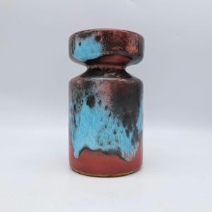 Könnte beinhalten: Eine Keramikvase mit zylindrischem Körper und ausgestellter Oberseite. Die Vase hat ein Farbschema aus Rot, Schwarz und Türkisblau. Die Glasur hat einen gesprenkelten, getropften Effekt, der der Oberfläche Textur verleiht. Die Vase ist ein dekorativer Gegenstand.